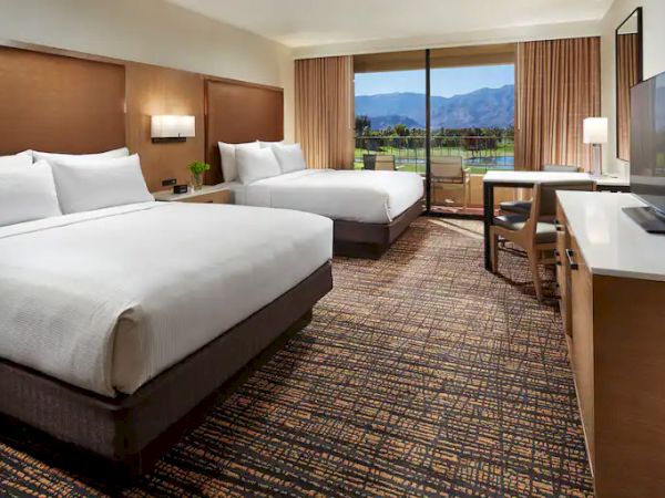 A hotel room with two beds, a TV, desk, and large window showing a scenic view of mountains and greenery.