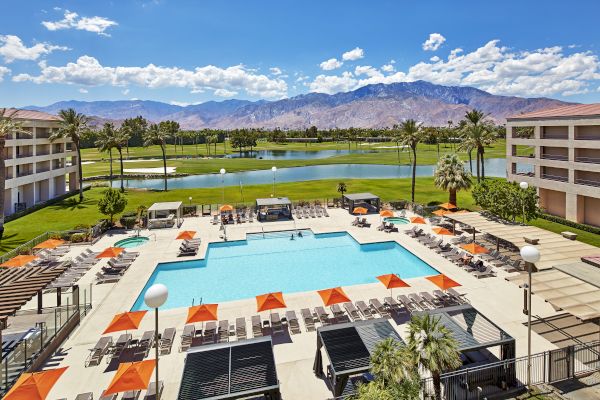 The image shows a resort pool area with orange umbrellas, surrounded by lounge chairs, buildings, a golf course, and mountains in the background.