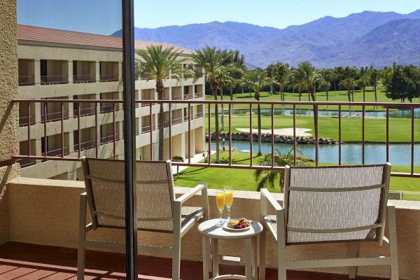 A balcony view with two chairs, a small table with drinks, overlooking a golf course, palm trees, and mountains in the background.