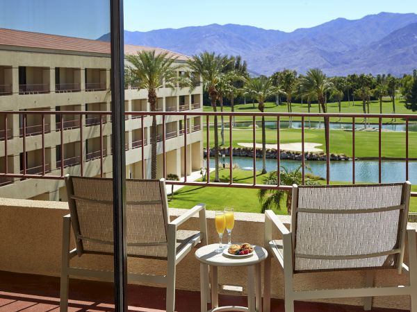 A balcony view with two chairs, a small table with drinks, overlooking a golf course, palm trees, and mountains in the background.