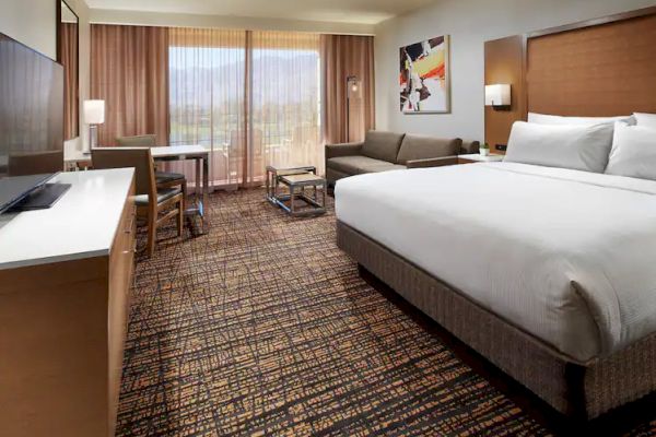 A hotel room with a king-size bed, sofa, TV, desk, and large window with curtains, showcasing a scenic mountain view.