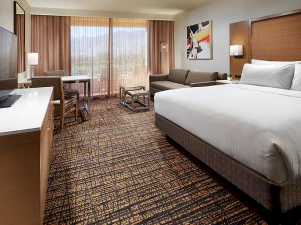 A hotel room with a king-size bed, sofa, TV, desk, and large window with curtains, showcasing a scenic mountain view.
