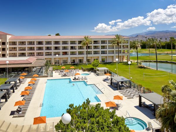 A hotel with a large swimming pool, lounge chairs, orange umbrellas, palm trees, and a scenic view of mountains and a manicured lawn.