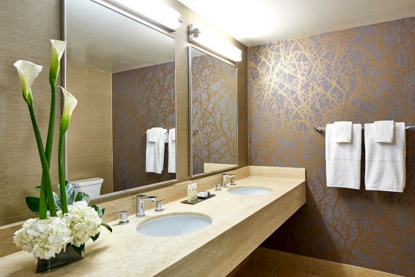 A modern bathroom with a large mirror, dual sinks, floral decorations, and towels on a textured wall.