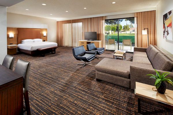The image shows a spacious hotel room with a bed, sofa, chairs, desk, TV, and large window with curtains opening to a view outside.
