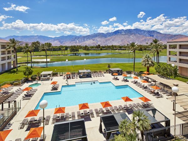 A resort with a swimming pool, orange umbrellas, and lounge chairs, surrounded by buildings, golf course, palm trees, and mountains in view.