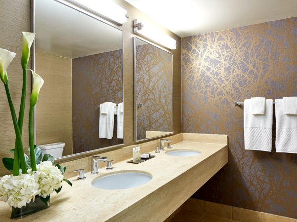 A modern bathroom with a double vanity, elegant lighting, decorative wallpaper, flowers, and neatly hung towels on the wall.
