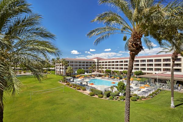 The image shows a hotel with a pool area, surrounded by palm trees and a lawn under a clear blue sky.