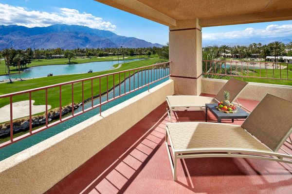 A patio with lounge chairs overlooks a golf course scene with a pond, greenery, and distant mountains under a blue sky.