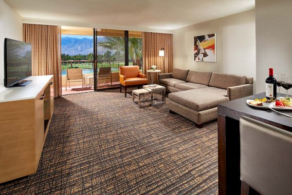 The image shows a modern hotel room with a sofa, TV, and a view of a terrace overlooking a golf course and mountains in the background.