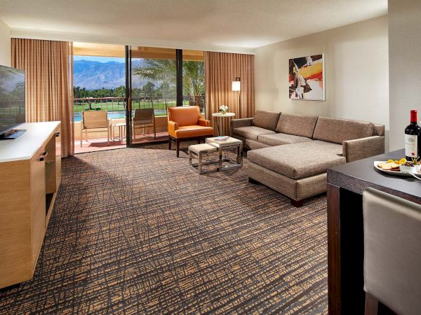 The image shows a modern hotel room with a sofa, TV, and a view of a terrace overlooking a golf course and mountains in the background.