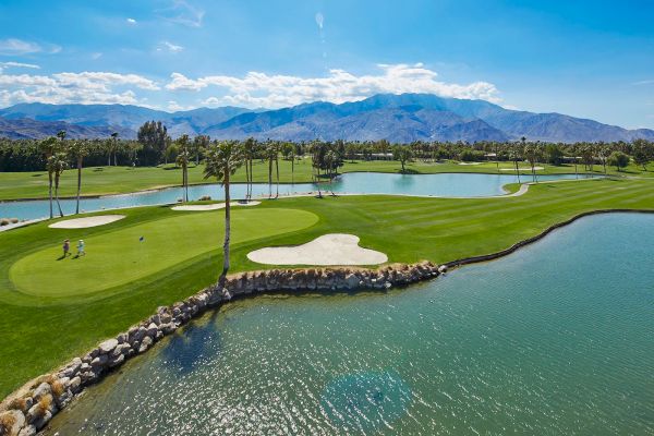 A golf course with water features, palm trees, and mountains in the background under a clear blue sky.