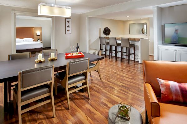 The image shows a modern living and dining area with hardwood floors, a dining table, a TV, and a bar counter with stools.