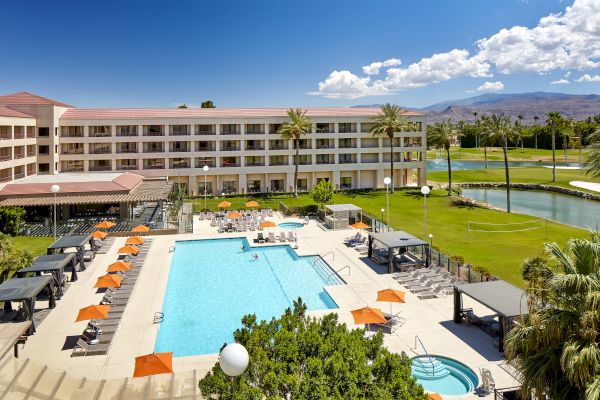 A hotel with a large pool, sun loungers, orange umbrellas, and palm trees, set against a mountainous backdrop.
