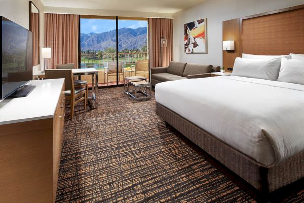 A hotel room with a large bed, TV, sofa, and table, overlooking a balcony with mountain views through a glass door.