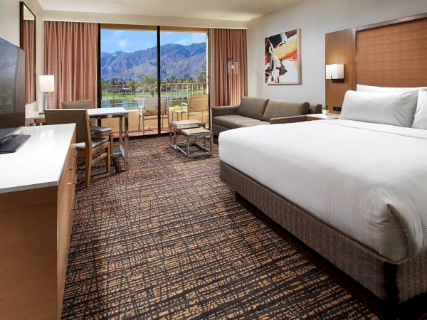 A hotel room with a large bed, TV, sofa, and table, overlooking a balcony with mountain views through a glass door.