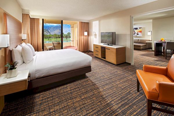 The image shows a spacious hotel room with a bed, TV, armchair, and a view of mountains through a window, leading to a sitting area.