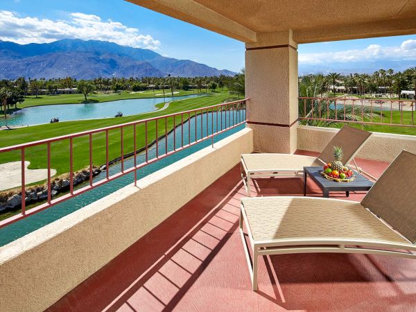 A balcony with lounge chairs overlooks a golf course and lake, offering a view of mountains under a blue sky with clouds.