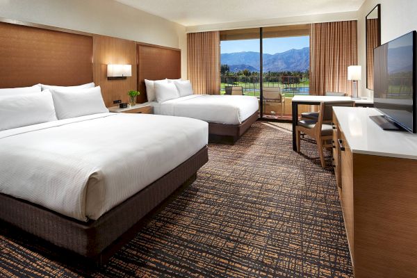 The image shows a hotel room with two large beds, a TV, a desk, and a view of mountains through a large window with natural light.