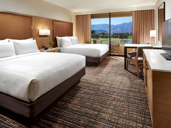 A modern hotel room with two large beds, a TV, desk, and a window view of mountains and nature.