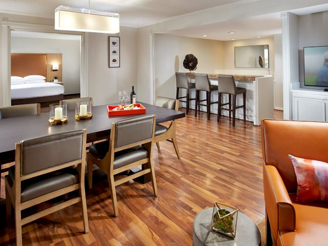 A modern living area features a dining table, bar seating, flat-screen TV, and a partial view of a bedroom and kitchen.