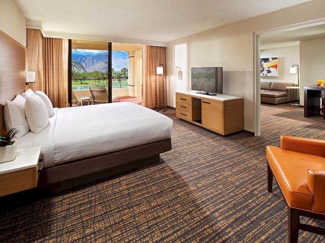 The image shows a modern hotel room with a large bed, TV, chair, and view of mountains. There’s also a separate seating area visible.
