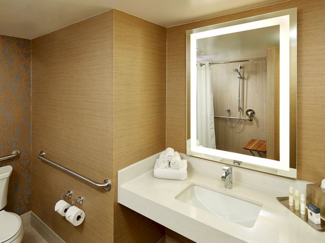 The image shows a modern bathroom with a sink, illuminated mirror, toilet, and accessible shower area with a bench.