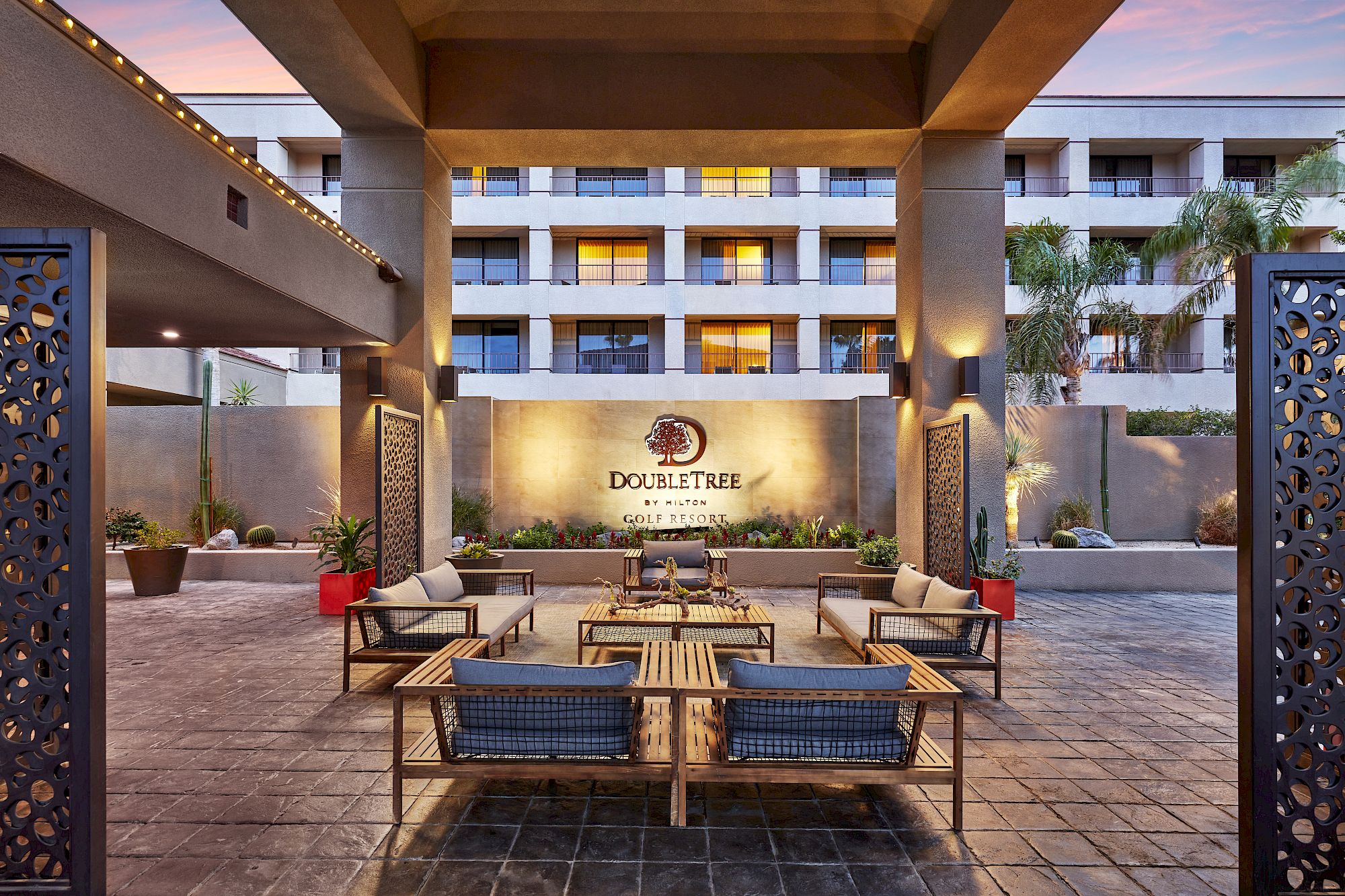 The image shows an outdoor seating area of a hotel, featuring wooden furniture and decorative panels, with the hotel building in the background.