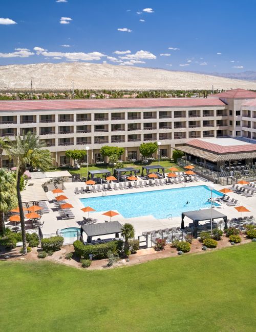 A large hotel with a central courtyard featuring a swimming pool, surrounded by lounge chairs with orange umbrellas, and palm trees.