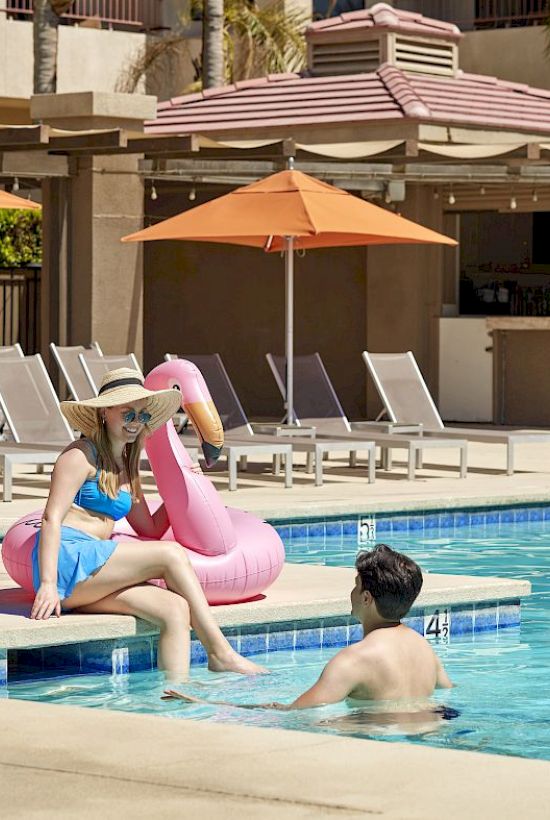 A woman in a swimsuit sits by a pool on a pink flamingo float, talking to a man in the water. Umbrellas and lounge chairs in the background.