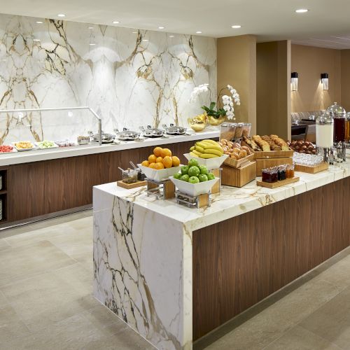 The image shows a buffet setup with fruits, pastries, and beverages on marble counters in an elegant setting.