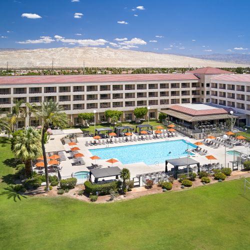 A hotel with a courtyard features a large pool, orange umbrellas, palm trees, and a volleyball net, set against a mountainous backdrop.