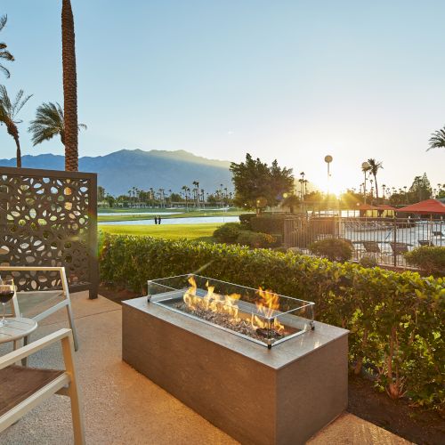 A patio with two chairs overlooks a pool, golf course, and mountains. A fire pit is in the center, with drinks on the table nearby.