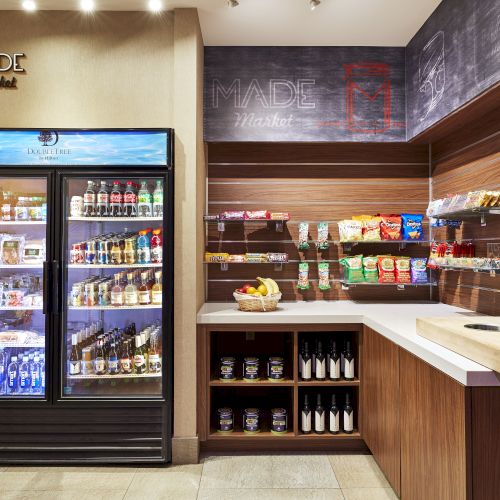 The image shows a small market corner with refrigerated drinks, snacks, wine bottles, a coffee chalkboard, and a light wooden counter.