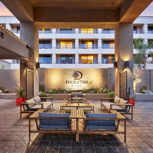 The image shows an outdoor seating area at a hotel entrance, with a modern design and decorative elements, under a clear sky.