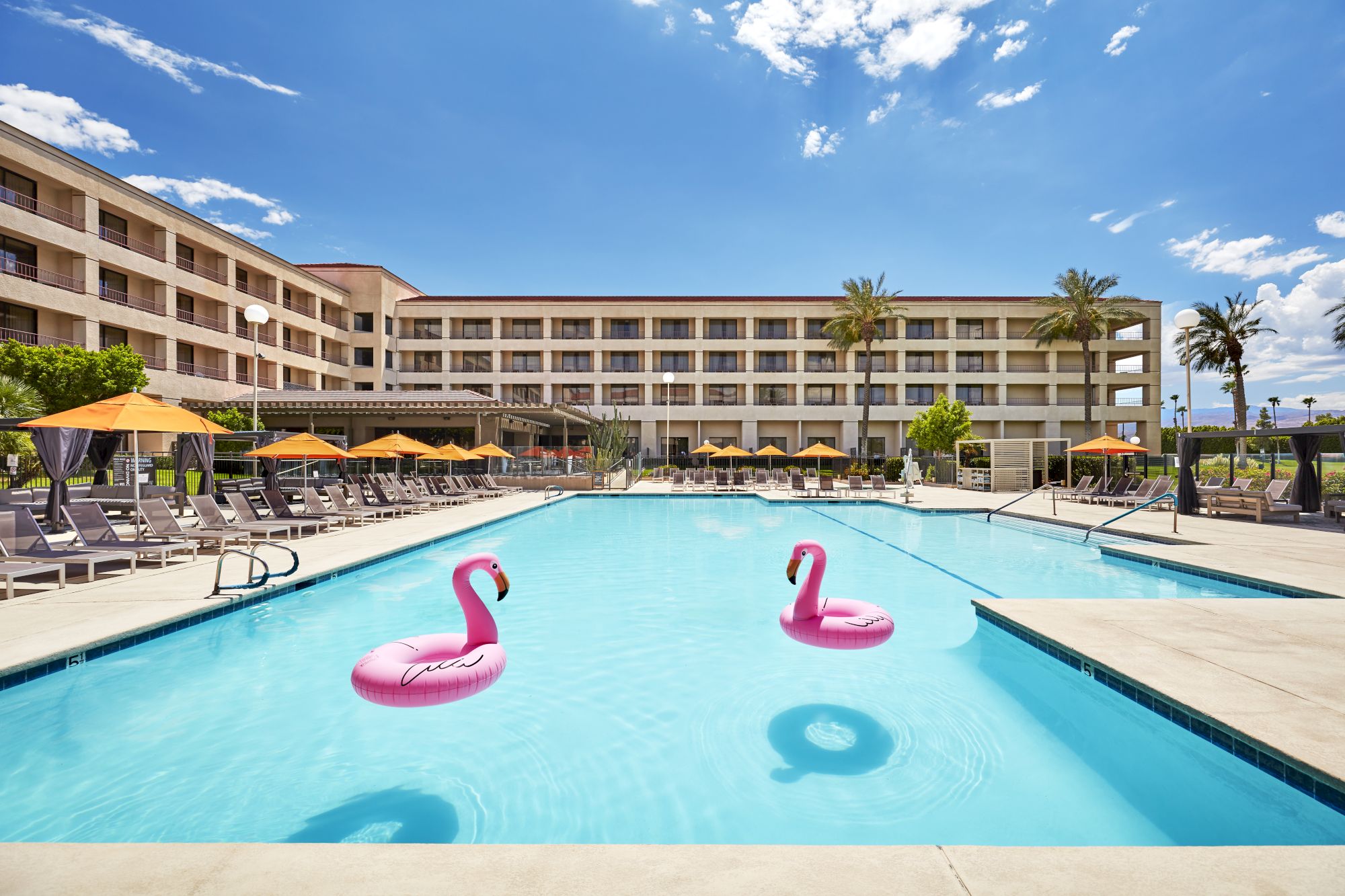 A hotel pool with pink flamingo floats, surrounded by lounge chairs and umbrellas, under a clear blue sky.