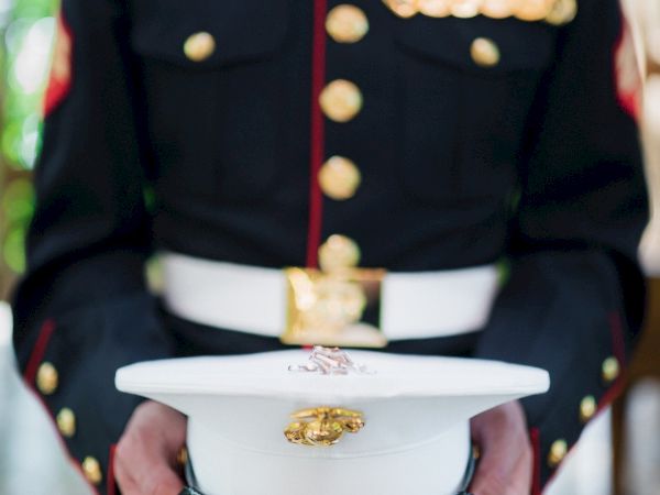 A person in a military dress uniform holds a white cap adorned with an emblem and medals on the jacket.