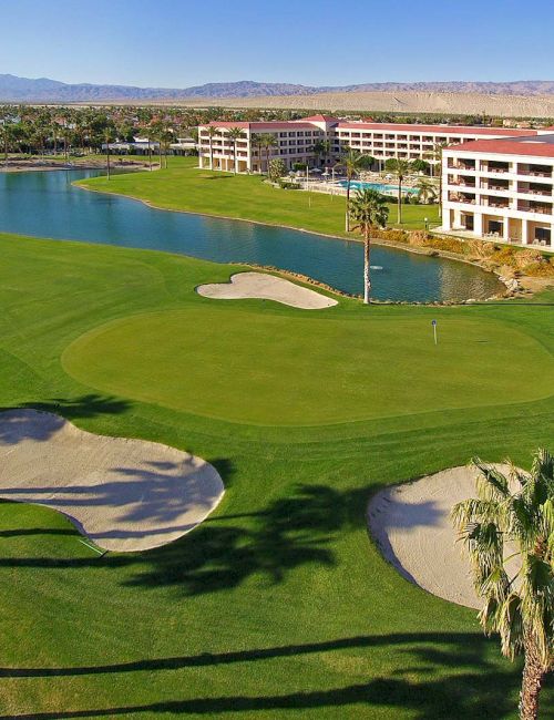 A lush golf course with sand bunkers, a pond, palm trees, and buildings in the background under a clear blue sky.