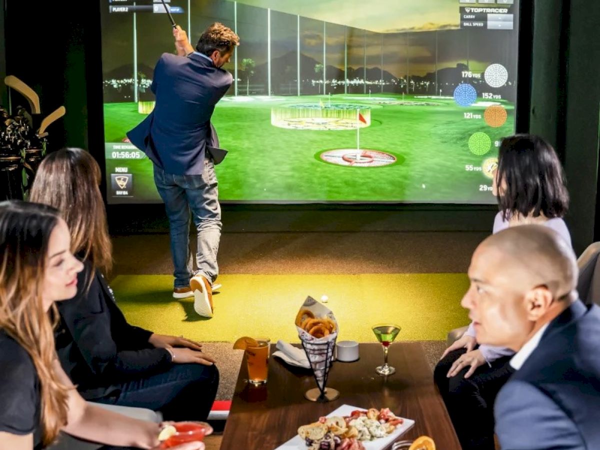 People enjoying a golf simulator in a stylish indoor setting, with snacks and drinks on the table.