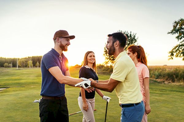 Four people are on a golf course, shaking hands and smiling, with golf clubs visible and the sun setting in the background.