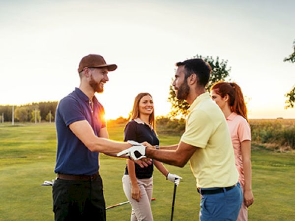 Four people are on a golf course, shaking hands and smiling, with golf clubs visible and the sun setting in the background.