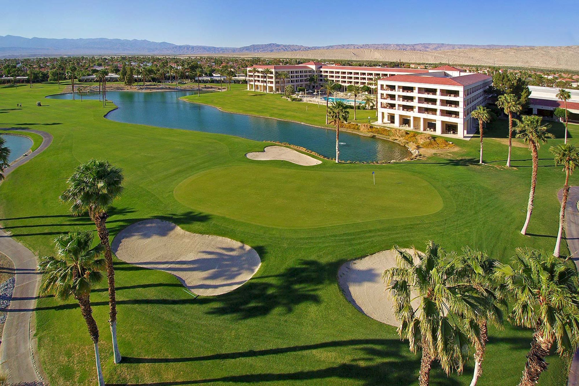 The image shows a golf course with sand traps, a pond, palm trees, and a nearby resort building. Mountains are visible in the background.