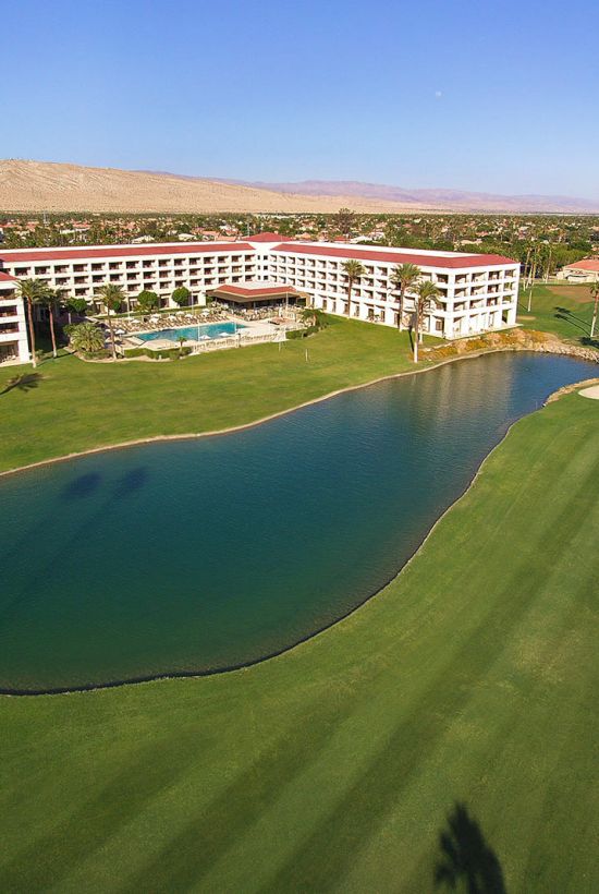 Aerial view of a resort with a large hotel building, surrounded by a golf course, water features, and palm trees under a clear sky.
