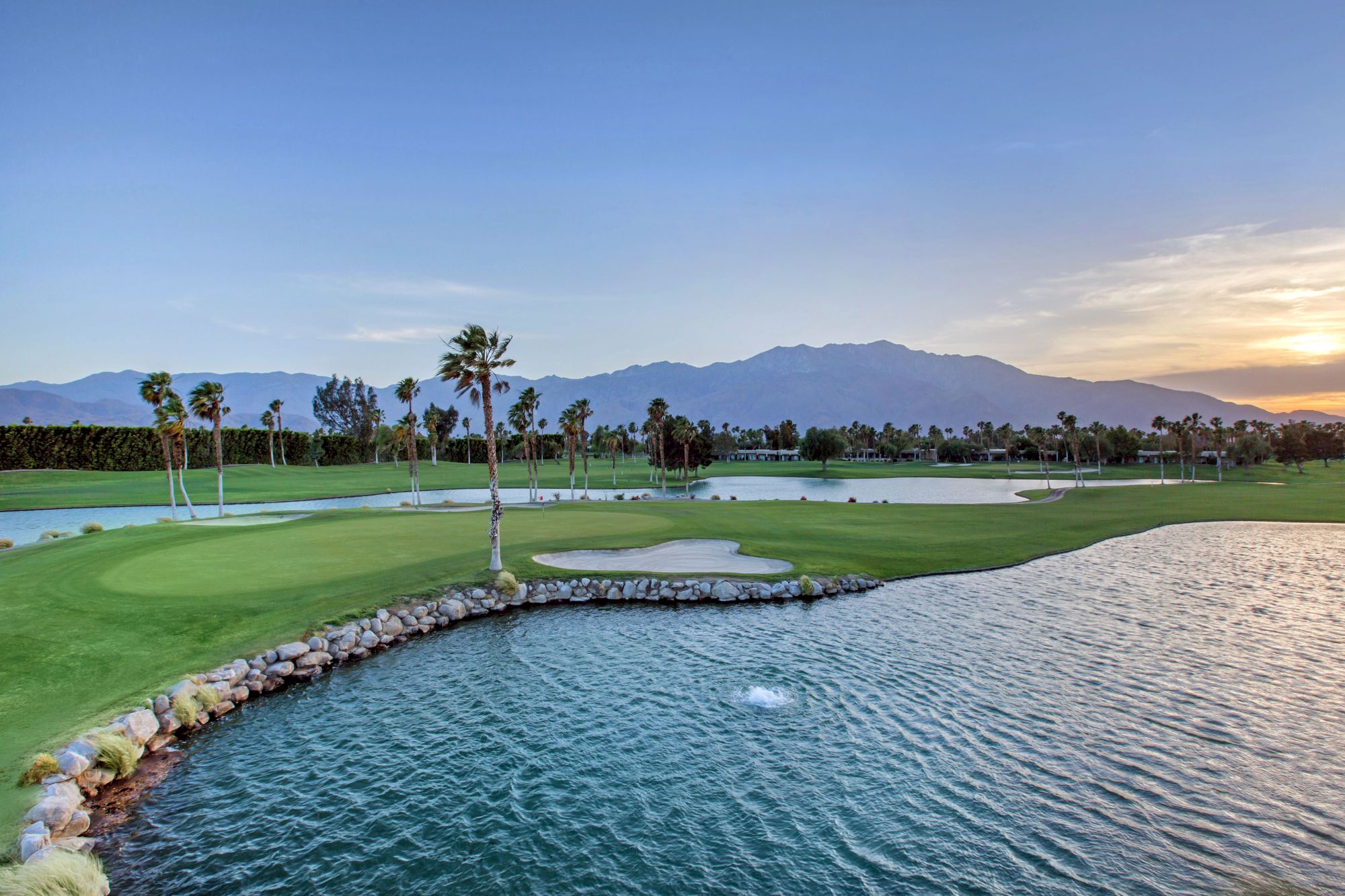 A serene golf course at sunset with palm trees, a pond, and mountains in the background.