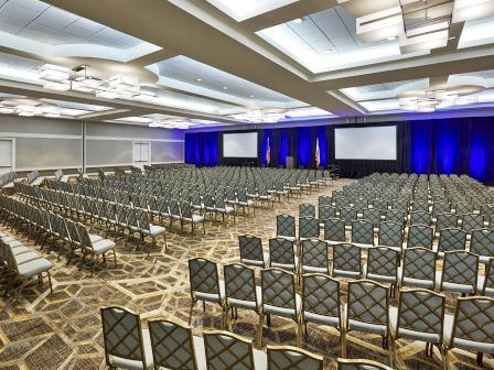 The image shows a large, empty conference room with rows of chairs, two projection screens, and blue lighting at the front.