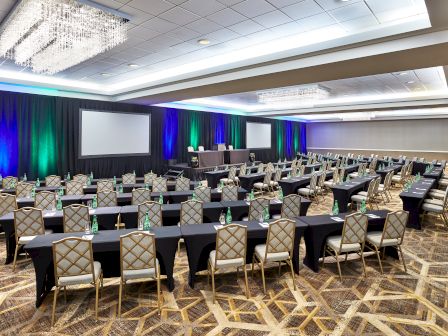 The image shows a large conference room with rows of tables, chairs, and a stage with screens, prepared for a formal event or presentation.