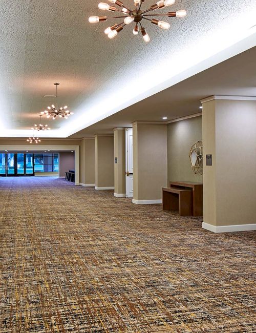 A spacious hallway with carpeted flooring, contemporary chandeliers, and open double doors leading to more rooms or a different area.
