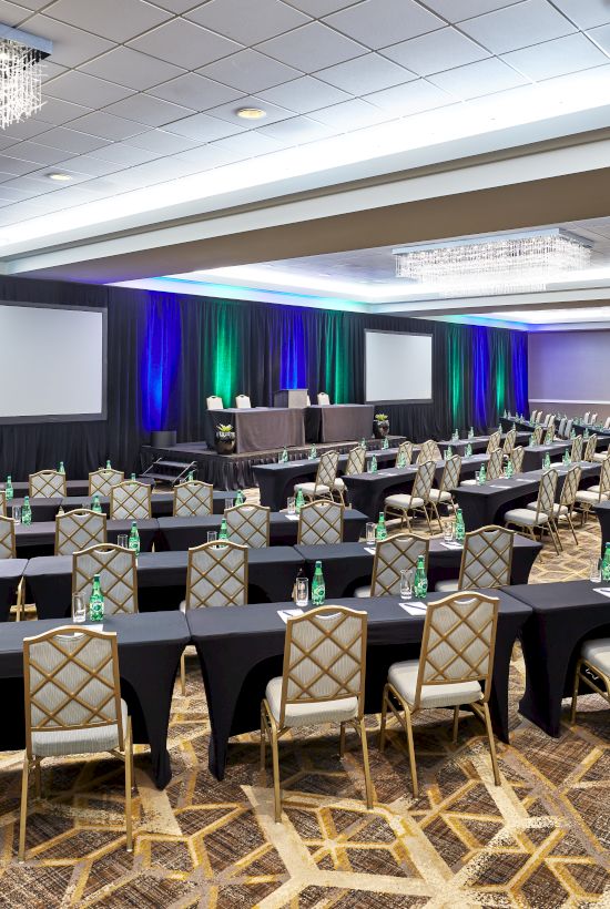 A conference room set up classroom-style with rows of tables and chairs, two projection screens, and a colorful backdrop with stage lighting.