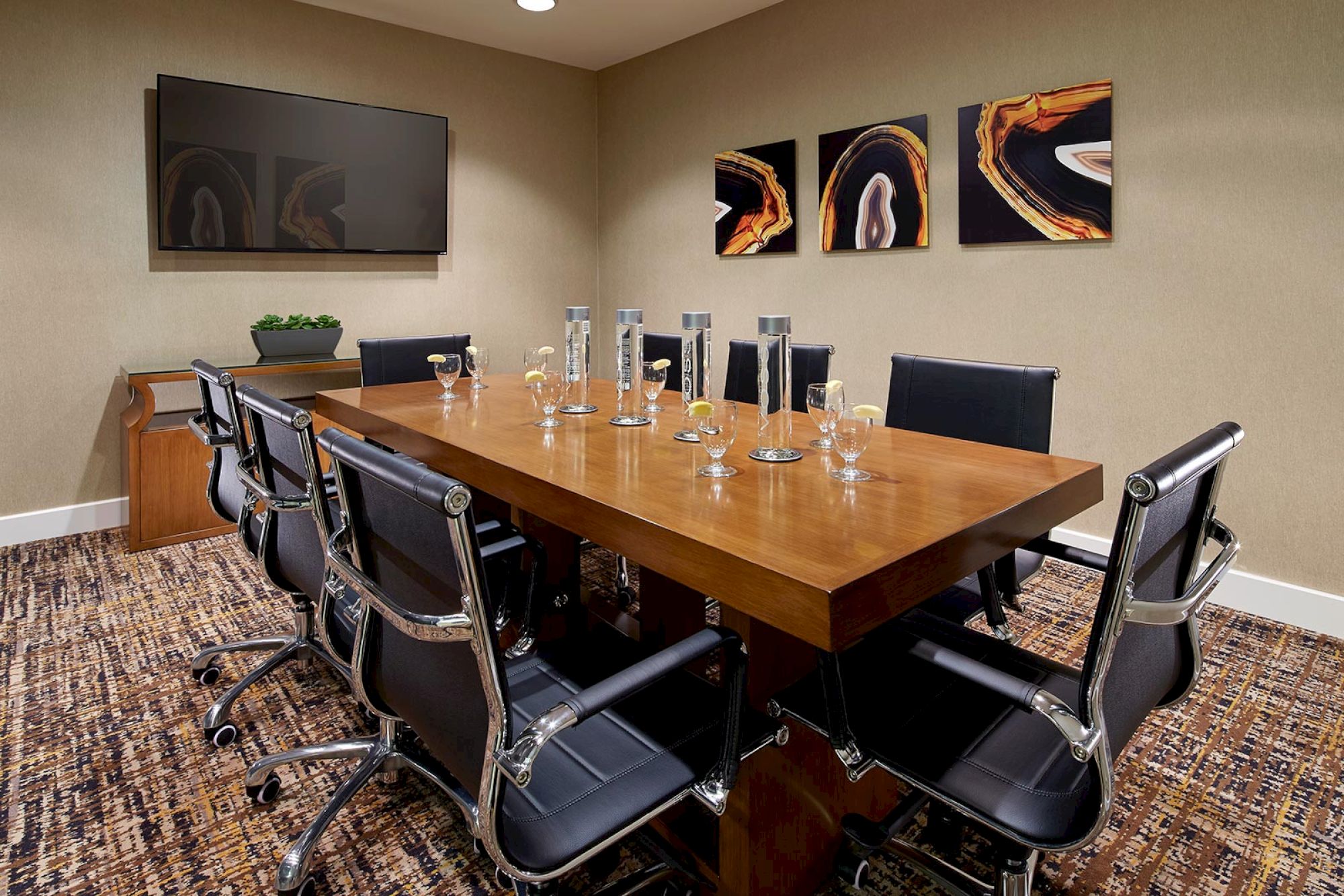 A conference room with a wooden table, eight chairs, a wall-mounted TV, abstract artwork, and water glasses is shown.