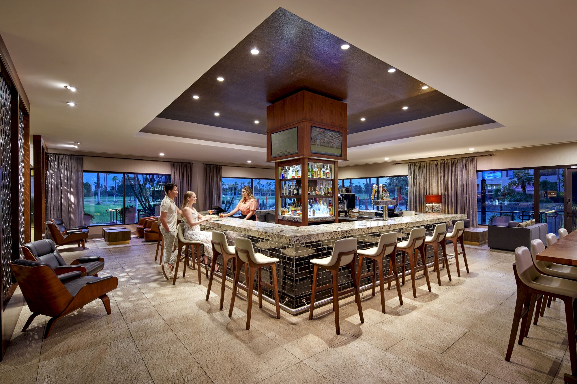 A modern bar with people seated around it, featuring stylish decor, ample seating, and large windows for natural light.